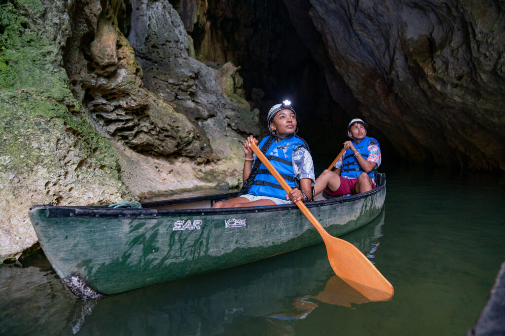A couple canoeing through a limestone cave in Belize, wearing helmets and life vests as they paddle along calm green water, one of the best nature experiences in Belize for couples.