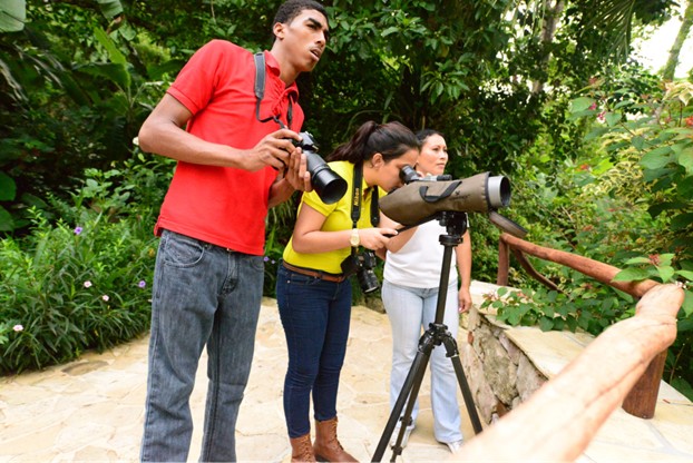 Three people birdwatching with binoculars and a spotting scope along a forest trail in Belize.
