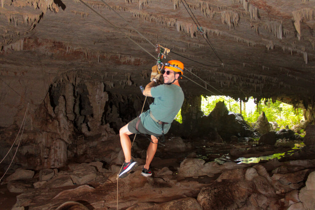 An aerial view of two adventurers wearing safety gear as they navigate a steep, rocky cliffside surrounded by dense jungle, a highlight of Belize's outdoor travel.